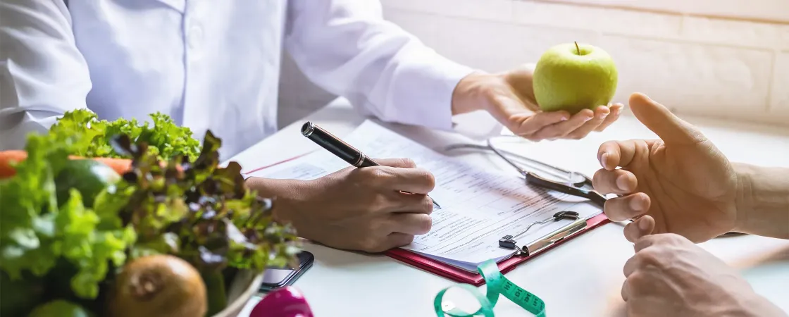 Doctor writing on pad and holding apple while advising patient.